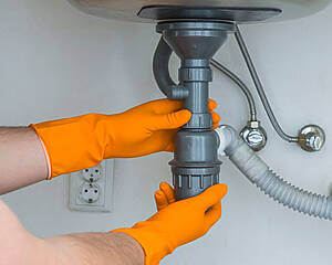 Plumber In Orange Rubber Gloves Clearing A Clogged Drain Under T