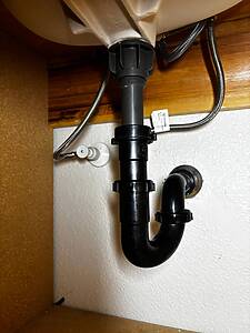Under-sink plumbing with a black P-trap and gray drain tailpiece in a cabinet. Braided metal supply lines run nearby.