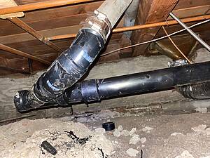 Basement ceiling with a large black insulated duct elbow and metal hose clamps crossing diagonally, with exposed copper pipes and wooden joists above.