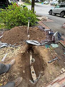 Open trench in front yard with exposed PVC pipe, a shovel, and a dirt pile; suburban street and cars in background.