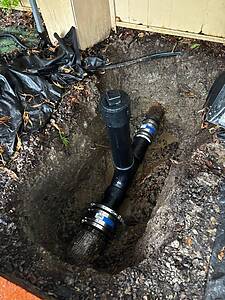Open trench beside a house showing black PVC pipes with a cleanout fitting and mud around the excavation area.