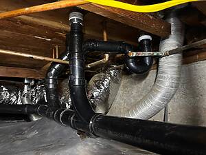 Underside of a basement ceiling showing black PVC drain pipes, copper water lines, and a metallic flexible duct in a damp crawl space.