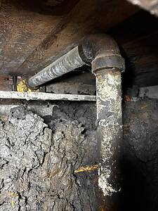 Underground plumbing pipe with elbow joint in a dark, dusty basement crawl space. A vertical pipe connects to a horizontal pipe under a wooden beam.