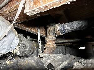 Dusty attic showing a rusted pipe, metal straps, and scattered debris among wooden beams and insulation dust.