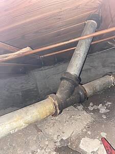 Exposed plumbing in a basement ceiling: a large gray PVC pipe and copper water lines running along wooden joists.