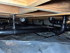Underside of a wooden floor in a crawl space with exposed pipes, ducts, and a plastic vapor barrier on the ground.