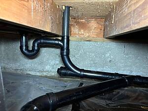 Exposed black plastic plumbing pipes running horizontally and vertically under a wooden floor joist in a crawl space, with cobwebs and dust.