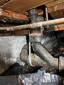 Cast-iron plumbing stack with a horizontal copper supply line in a dusty crawl space under a wooden floor.