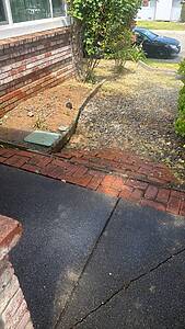 Driveway area with a brick border, gravel path, and a black car parked on the street in the background, beside a brick wall and greenery to the left