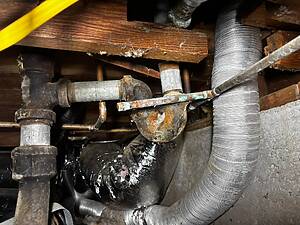 Cluttered crawl space with rusty pipes, exposed joists, and a large silver duct running vertically.