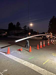 Two men at a night-time street worksite move a long white PVC pipe among orange traffic cones under a dark sky with a bright moon above houses and trees.