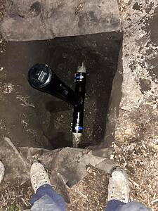 Person standing at the edge of a dirt shaft looking down at a black pipe entering the opening; muddy walls and concrete edges surround the shaft.
