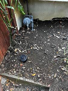 Gas meter and piping attached to a stucco wall in a bare dirt yard with fallen leaves nearby.