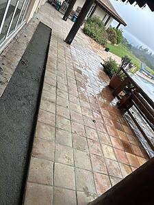 Wet terracotta tile patio along a house with wooden posts and potted plants, overlooking a grassy yard under a cloudy sky.