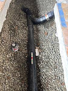 Black plastic drainage pipe with a curved junction resting on a bed of gravel beside a concrete wall.