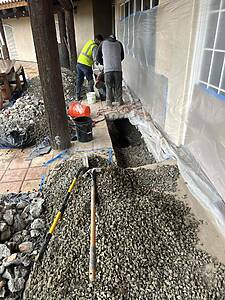 Construction workers in a yard dig a trench along a house exterior, with plastic sheeting taped to the wall and scattered gravel and tools nearby.