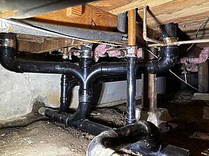 Basement plumbing view showing black PVC drain pipes, copper water lines, and wooden joists overhead.