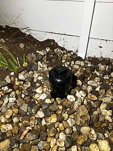Ground-level view of a garden bed with dark soil, mixed pebbles, and a black irrigation sprinkler head near a white wall.
