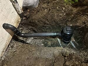 Exposed soil in a trench beside a concrete wall with black PVC pipes and a valve fitting visible at ground level.