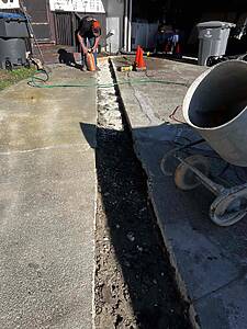 Man in a black shirt and orange cap digging a trench along a concrete driveway, bag of material in hand, with orange safety cones nearby.