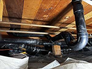 Pipes and wooden ceiling joists in a basement with black PVC plumbing running across and scraps of insulation below.