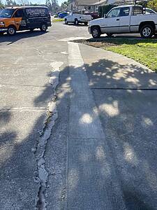 Residential street with a long concrete trench along a crack in the road, cars parked on both sides and a service van in the background after roadwork