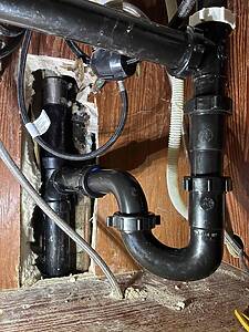 Undersink view of a black plastic P-trap connected to PVC drain pipes with braided and corrugated hoses in a wooden cabinet.
