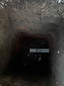 Deep, dark vertical shaft or trench in the ground with a light-colored pipe running along the bottom edge of the opening