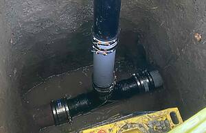 Vertical black underground pipe with a capped top standing in a dirt trench, connected to a lower horizontal pipe with orange clamps nearby.