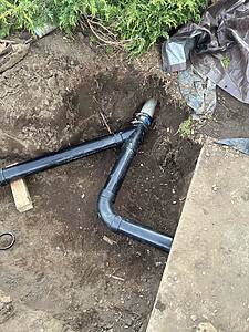 Digging a trench for underground plumbing with black PVC pipes connected by a joint, dirt all around and a tarp nearby.