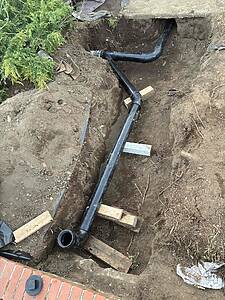 Trench with black PVC drainage pipe and elbow joints laid for installation, supported by wooden blocks amid dirt and a brick edge nearby.
