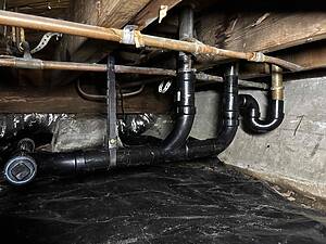 Basement crawl space with copper water pipes, black plastic drain pipes, metal straps, and a tape measure nearby.