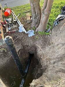 Open excavated trench beside a tree with two large black PVC pipes entering the ground, and scattered construction tools and a red bucket nearby in a yard.