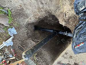Open trench with a large black PVC pipe, dirt walls, and scattered tools and tarp at a construction site.