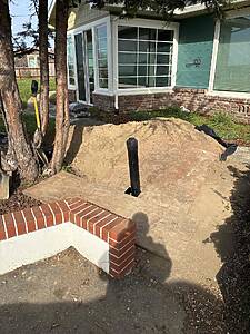 Front yard with a large dirt pile, a shovel, and a brick-bordered planter outside a teal house window.