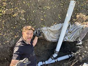 Man wearing a black polo and gloves in a muddy trench, posing for a selfie with blue PVC pipes laid out in the ground behind him.