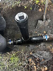 Trench with a black PVC pipe fitting and a metal inspection cap visible above mud and soil. A shovel lies nearby in damp ground.
