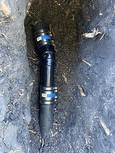Black plastic PVC pipe with blue labeling lying in a muddy trench, part of an underground drainage or irrigation line.