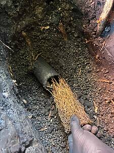 Person wearing gloves looks into a dark, muddy excavation with a broken black pipe and tangled straw-like fibers nearby.