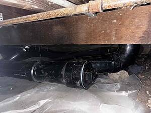 Underside view of a dusty crawl space showing black PVC drain pipes beneath a wooden beam with a rusted metal pipe above.