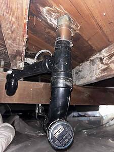 Underside of a sink with black PVC drain pipes, a copper coupling, and wooden joists nearby.