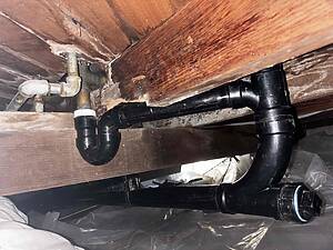 Exposed plumbing under a wooden floor, showing PVC traps and metal fittings in a dim crawl space.