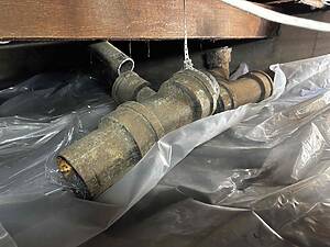 Corroded metal plumbing pipes under a wooden beam, secured with a perforated metal strap, with plastic sheeting beneath.