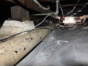 Low-angle view of a dusty crawl space with exposed pipes, wooden beams, and plastic sheeting on the floor behind a concrete footing.