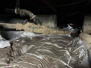 Rusty metal pipe elbow in a dark basement utility area, with plastic sheeting on the floor.