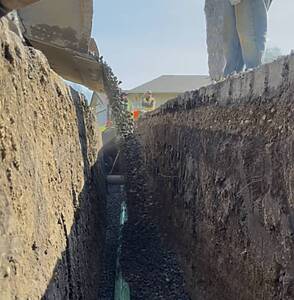 Trenched construction site with a green pipeline laid along gravel, workers standing at the far end under a clear sky.