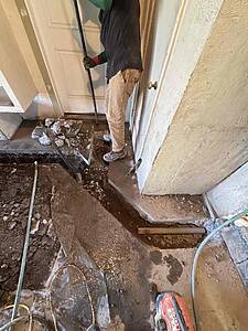 Construction worker in dirt-stained clothes stands by a doorway, holding a long tool over a trench filled with broken concrete and debris.