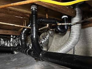 Underground-looking basement area with black plastic pipes, metal fittings, and a gray flexible duct visible among wooden joists.