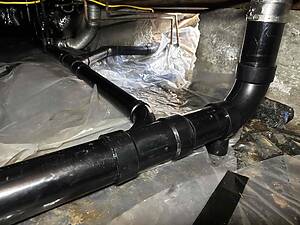Underside of a basement crawl space showing large black PVC pipes, foil insulation, and a concrete wall.