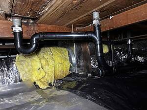 Underside of a wooden ceiling with black plastic drain pipes and metal couplings in a basement plumbing area, with yellow insulation below and foil wrap nearby.